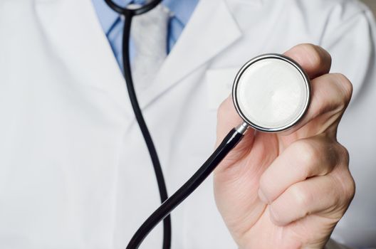 Close up of a Doctor's hand, holding a stethoscope outstretched towards the viewer.  Doctor's white coat provides space to the left side of frame.