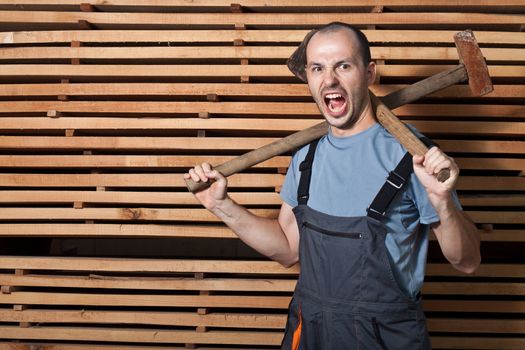 Angry worker with axe and hammer. Horizontal shot with wood in the background.