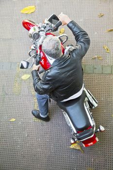 Diagonal aerial viewpoint of a stationary red motorbike and a Grey haired senior rider in leather jacket, denim jeans and boots on a tied floor with fallen autumn leaves. Generic shot taken in Bombay India