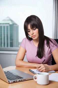 Beautiful business woman writing down some notes in notepad with laptop on desk