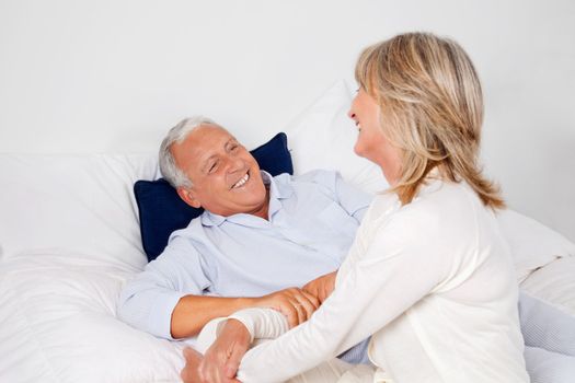 Happy senior man lying in bed with mature woman sitting beside him