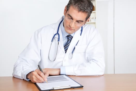 Male Doctor writing on clipboard sitting at desk.