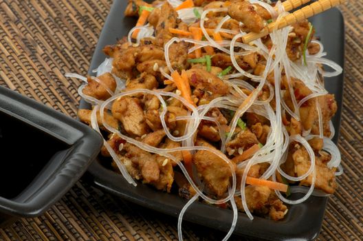 Picked up Piece of Chicken Teriyaki From Bowl by Pair of Chopsticks and Soy Sauce in Black Square Bowl closeup on Straw mat background