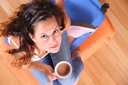 A young woman sitting on a stack of suitcases while drinking coffee and waiting for the departure to vacations.