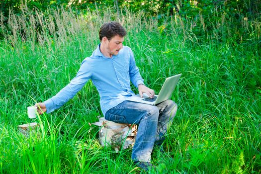 Young man outdoors with a laptop reaching his coffee