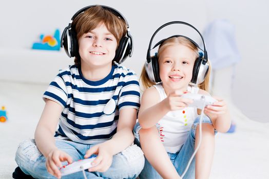 cute boy and girl playing gaming console in wireless headphones, sitting on the floor