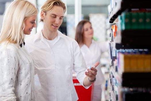 Pharmacist And Customers At Pharmacy In Front Of Shelves With Pharmaceuticals.