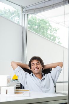 Happy young male architect looking at model house while sitting in chair with hands behind head