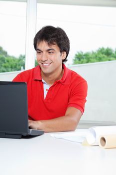 Young man smiling while working on laptop from home with rolled papers on desk