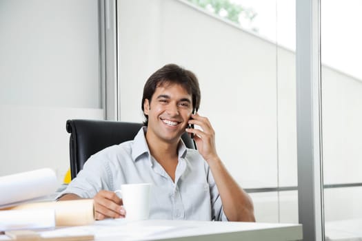 Portrait of cheerful young male architect answering phone call while having coffee