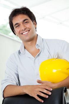 Portrait of happy young male architect holding yellow hardhat while standing by office chair