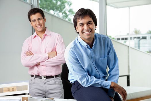 Portrait of young male architect smiling while colleague stands with arms crossed in background