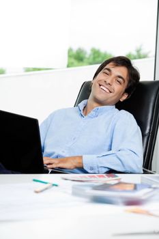 Portrait of happy young male interior designer with laptop sitting in office chair