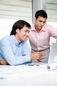 Young male architect pointing at laptop screen while colleague standing beside.