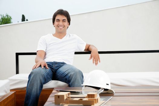 Portrait of a relaxed young male architect with hardhat and model structure on table