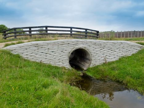 Large circular storm water culvert or drainage pipe passing under a scenic rural road with a rustic wooden fence above and concrete bank reinforcement and water below