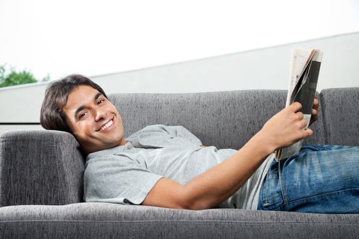 Portrait of young man in casual wear lying on sofa with a magazine