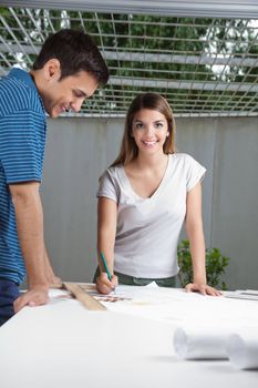 Portrait of young female architect working on blueprint with colleague standing by table