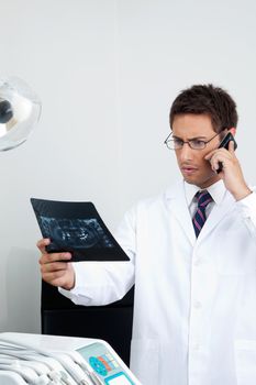 Young male doctor looking at X-ray report while using mobile phone in dental clinic