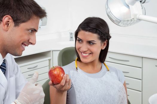 Happy male dentist showing thumbs up sign to a female patient holding an apple in clinic