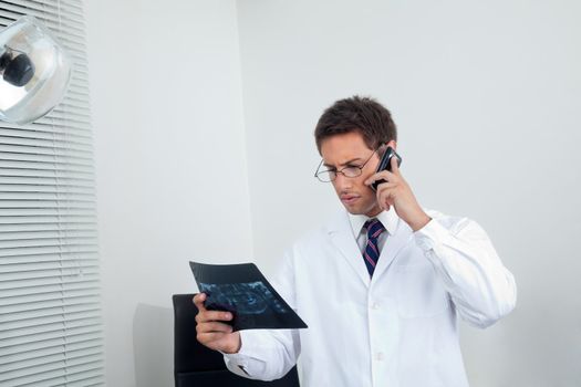 Young male dentist looking at X-ray report while using cellphone in clinic