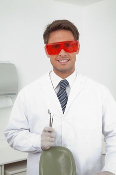 Portrait of happy young male dentist wearing protective eyewear while holding angled mirror and carver in clinic