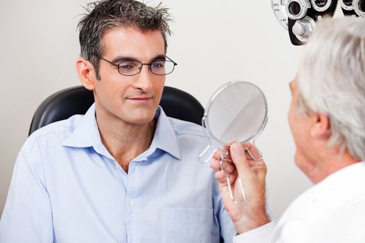Man in stylish reading glasses while looking at mirror with optometrist in the clinic