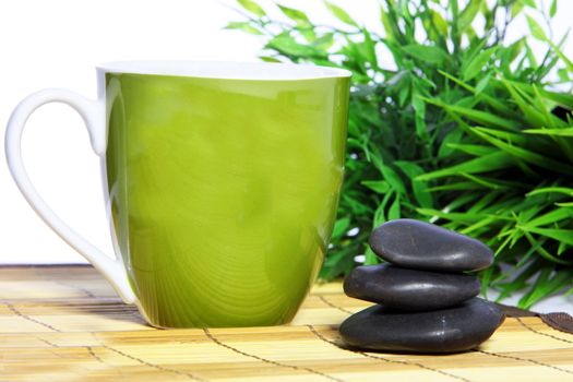 Pretty green glazed ceramic mug and spa massage stones standing on a reed mat in front of fresh green bamboo leaves in a spa, health and wellbeing concept