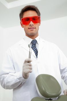 Portrait of young male dentist wearing protective eyewear while holding angled mirror and carver in clinic