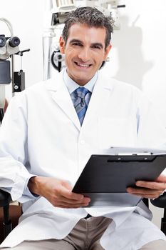 Confident optometrist smiling while holding a clipboard at his clinic