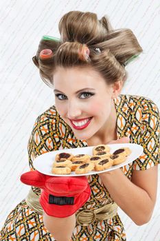 Portrait of pretty young woman with hair curlers holding plate of baked cookies