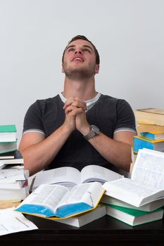 Young Student overwhelmed with studying with piles of books in front of him and praying