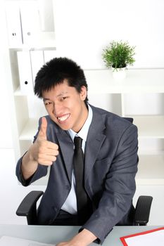 Smiling Asian businessman sitting at his desk in his office working giving a thumbs up of approval and success