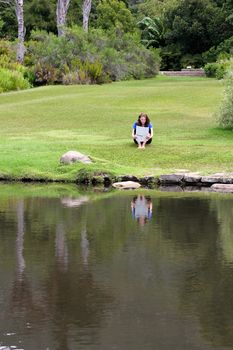 Woman reading newspaper sitting in grass with a pond in the foreground
