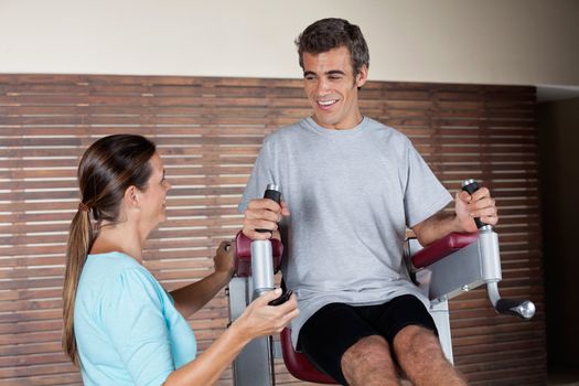 Happy young man using an exercise machine while looking at instructor in health club
