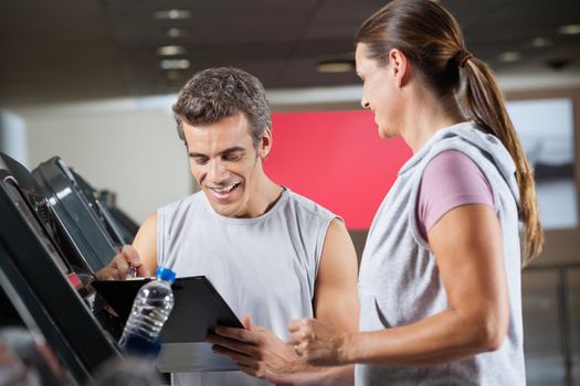 Happy male instructor holding clipboard while standing besides client exercising on treadmill in health club