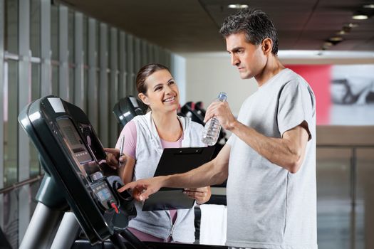 Happy female instructor looking at male client on treadmill drinking water in health center