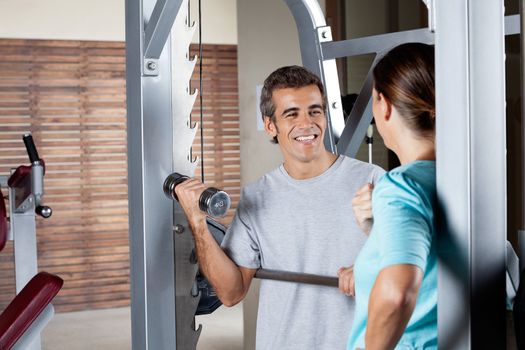 Happy young man lifting weights while looking at instructor at health club