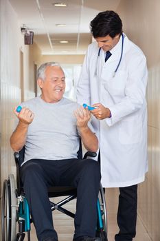 Happy young doctor giving hand weights to the senior man sitting in a wheelchair at hospital