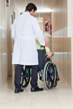 Rear view of a young doctor with senior woman sitting in wheel chair at hospital corridor