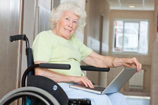 Happy senior woman sitting in wheelchair using laptop at hospital