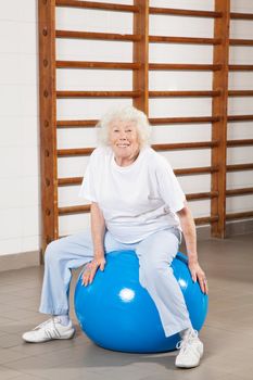 Full length portrait of a happy senior woman sitting on fitness ball at hospital gym