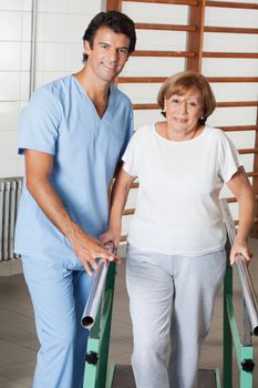 Portrait of a physical therapist assisting senior woman to walk with the support of bars at hospital gym