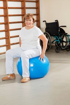 Full length portrait of a happy senior woman sitting on fitness ball at hospital gym