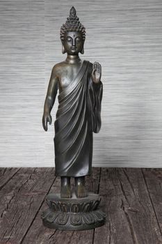 Metal statuette of Buddha standing in robes with his hand raised in a blessing on a wooden floor against a grey wall