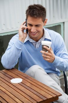 Handsome young man with disposable coffee cup communicating on cell phone while sitting at cafe