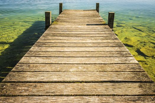 Jetty of weathered wood over a lake with blue water and green small waves and rocks at the bottom on a sunny day