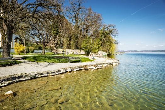 Shore of Lake Starnberg in Germany with gravel, trees, benches and people in sunny weather