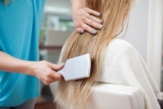 Beautician combing long blond hair of female customer with brush at parlor