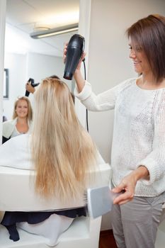 Pretty beautician blow drying female's long blond hair at parlor